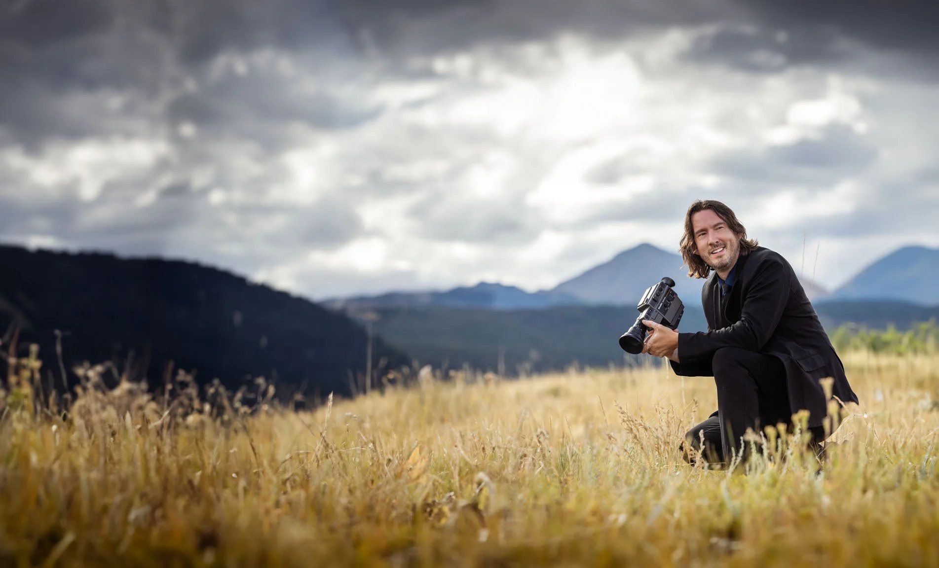 Adam Grumbo kneeling in a field of gold grass with a cinema camera, storm clouds and mountains behind him